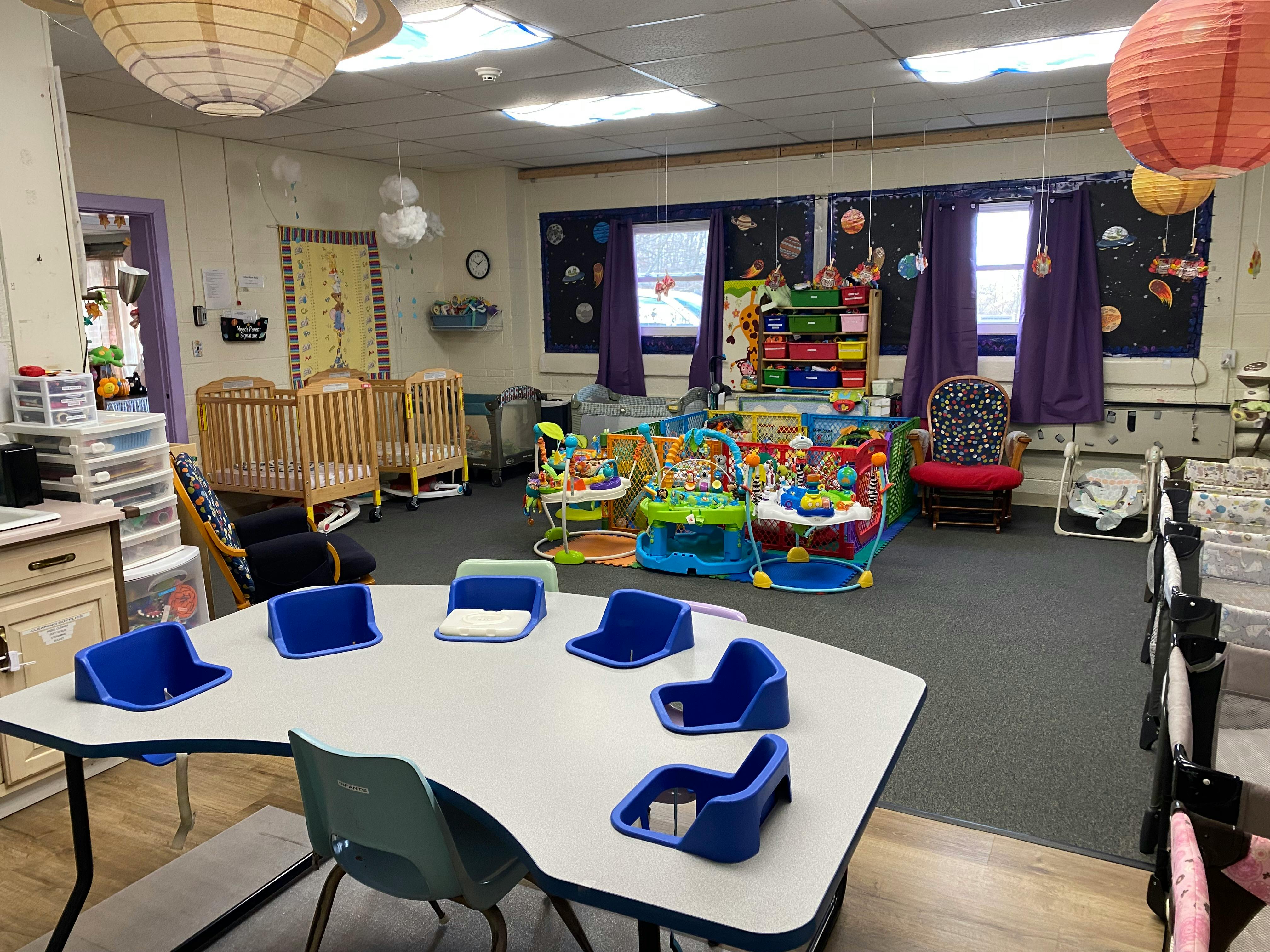 Classroom with small table and chairs, cribs, and infant activity centers
