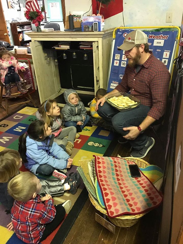 Person holding a tray of food, surrounded by children sitting on a rug