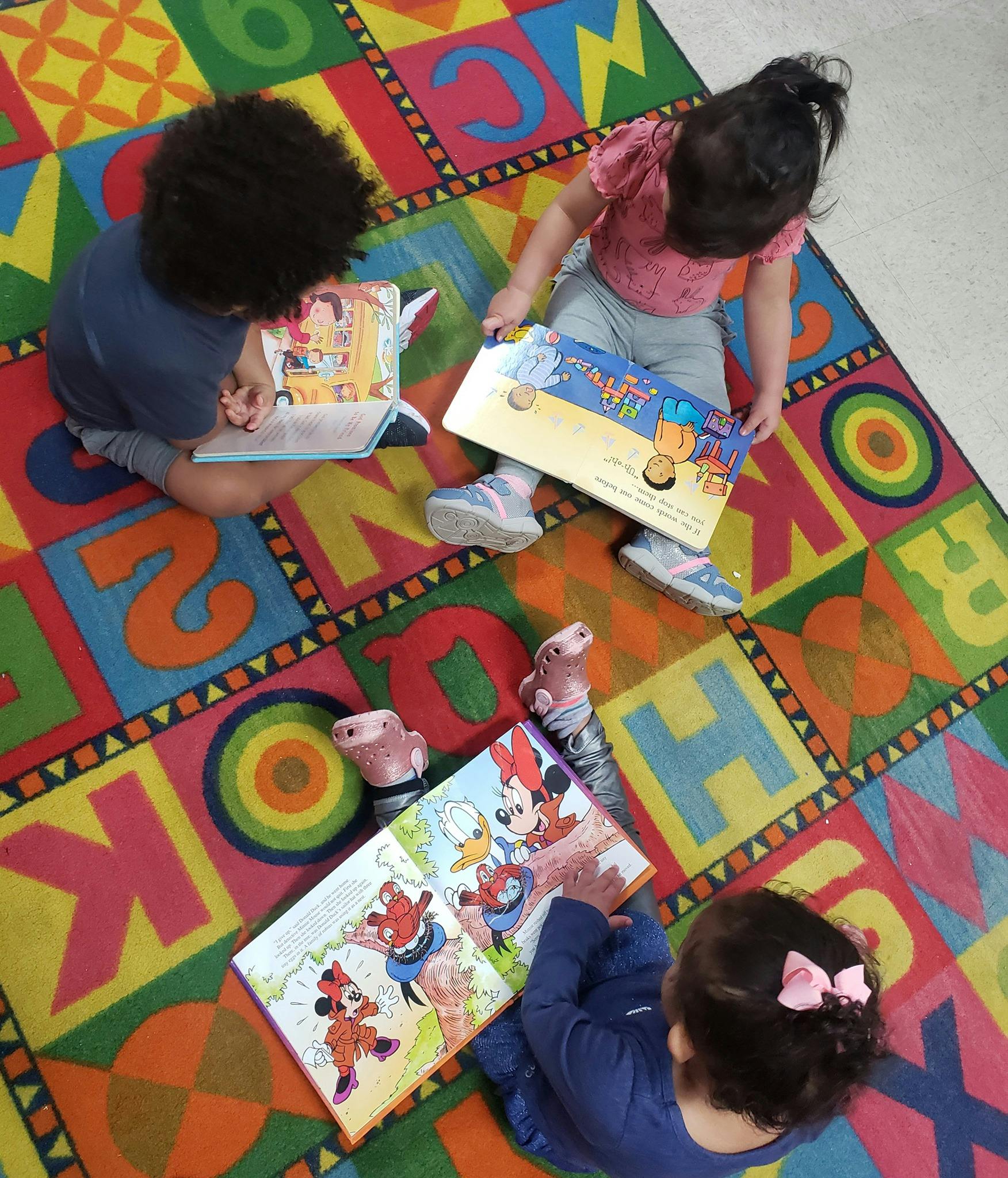 Three children sitting on a colorful alphabet rug, reading books