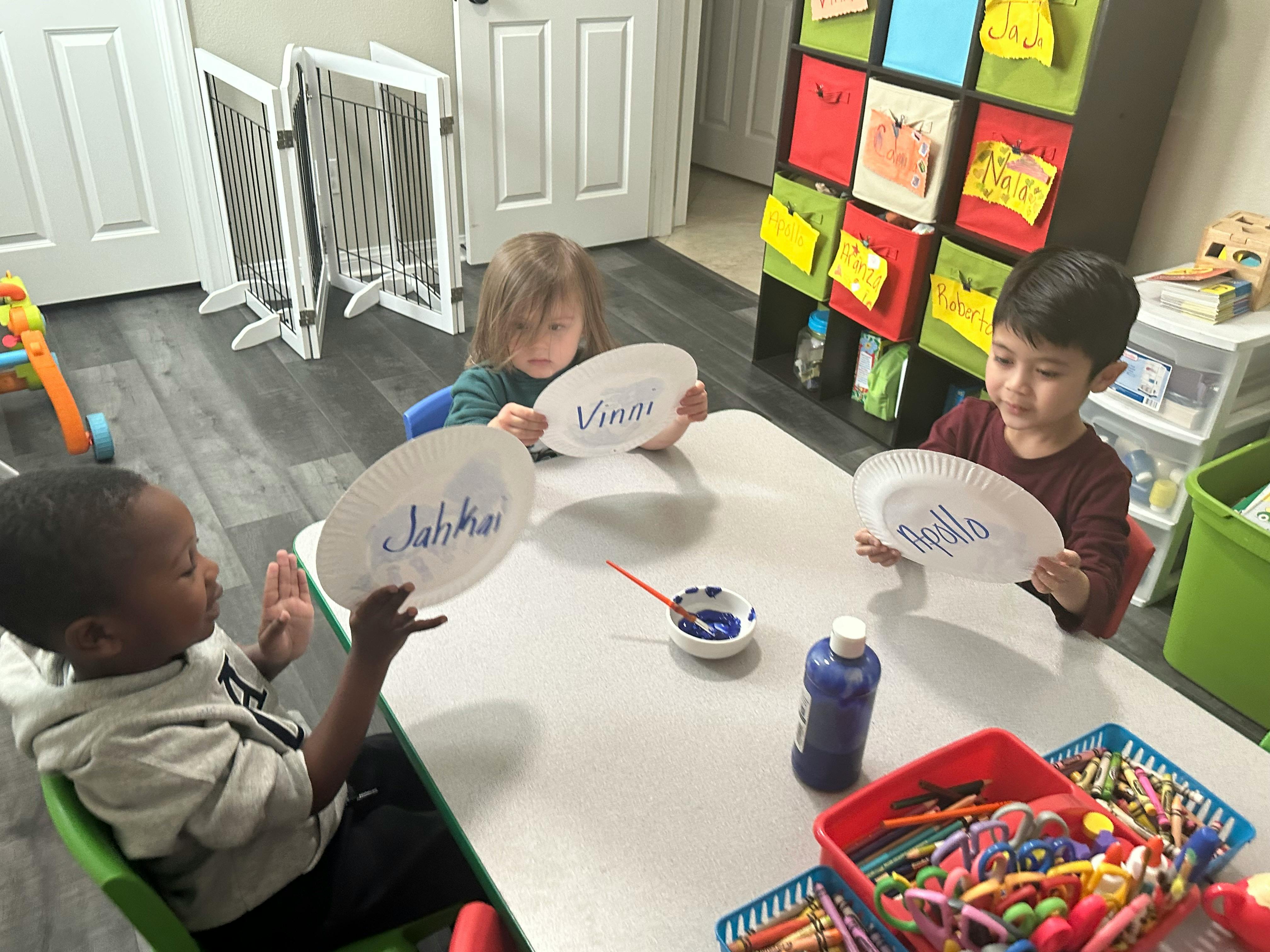 Three children holding paper plates with writing on them at a table