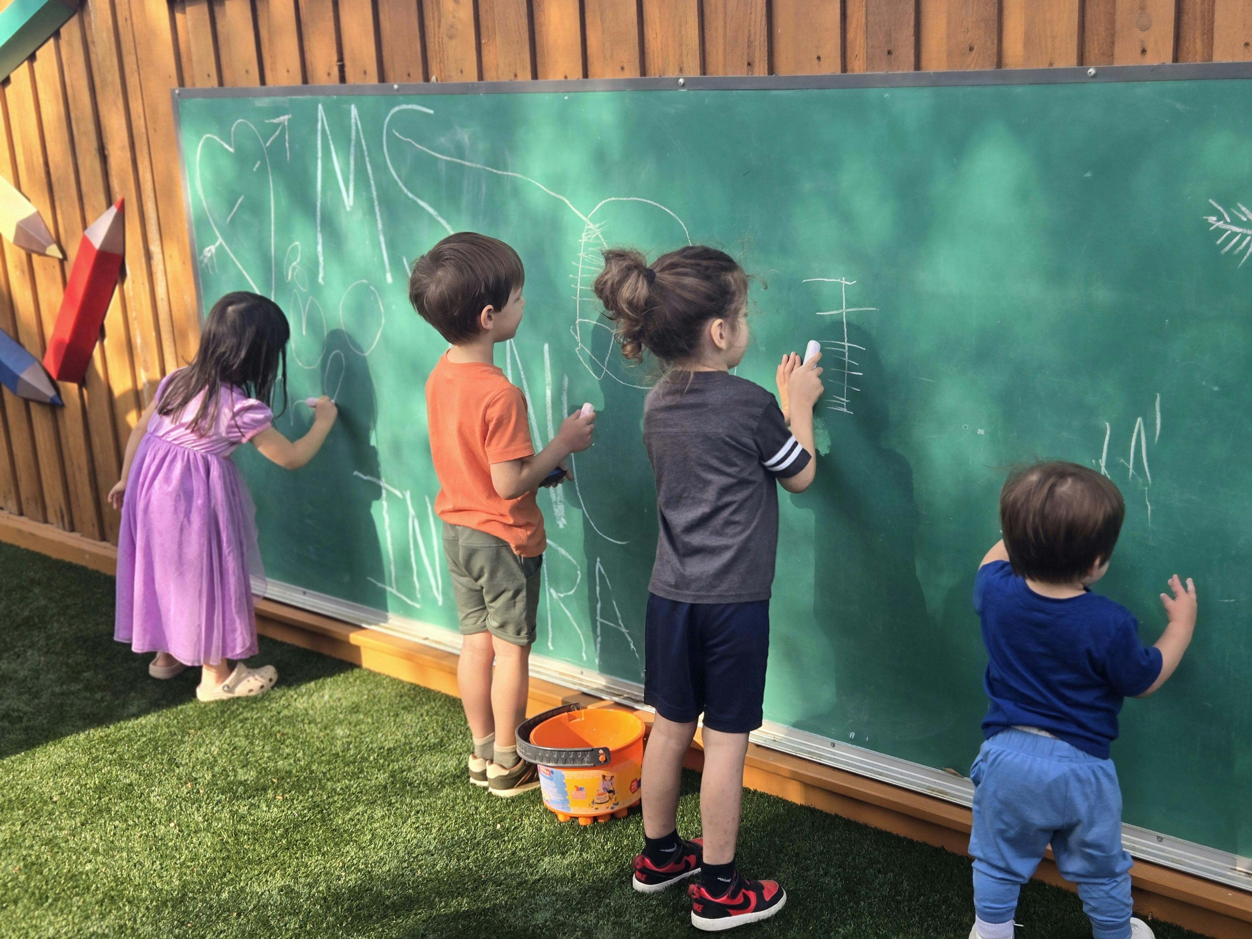 Four children drawing on a large green chalkboard outdoors