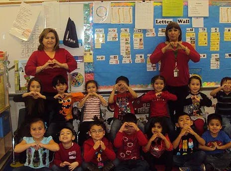 Two women and multiple children making heart shapes with their hands in a classroom