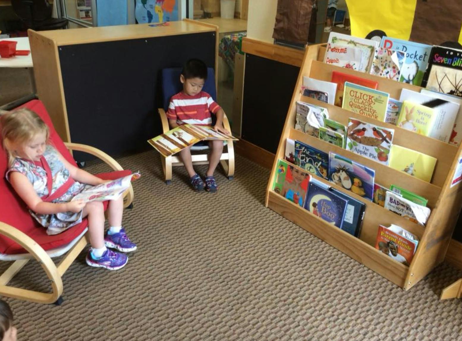 Two children sitting in chairs reading books in a classroom