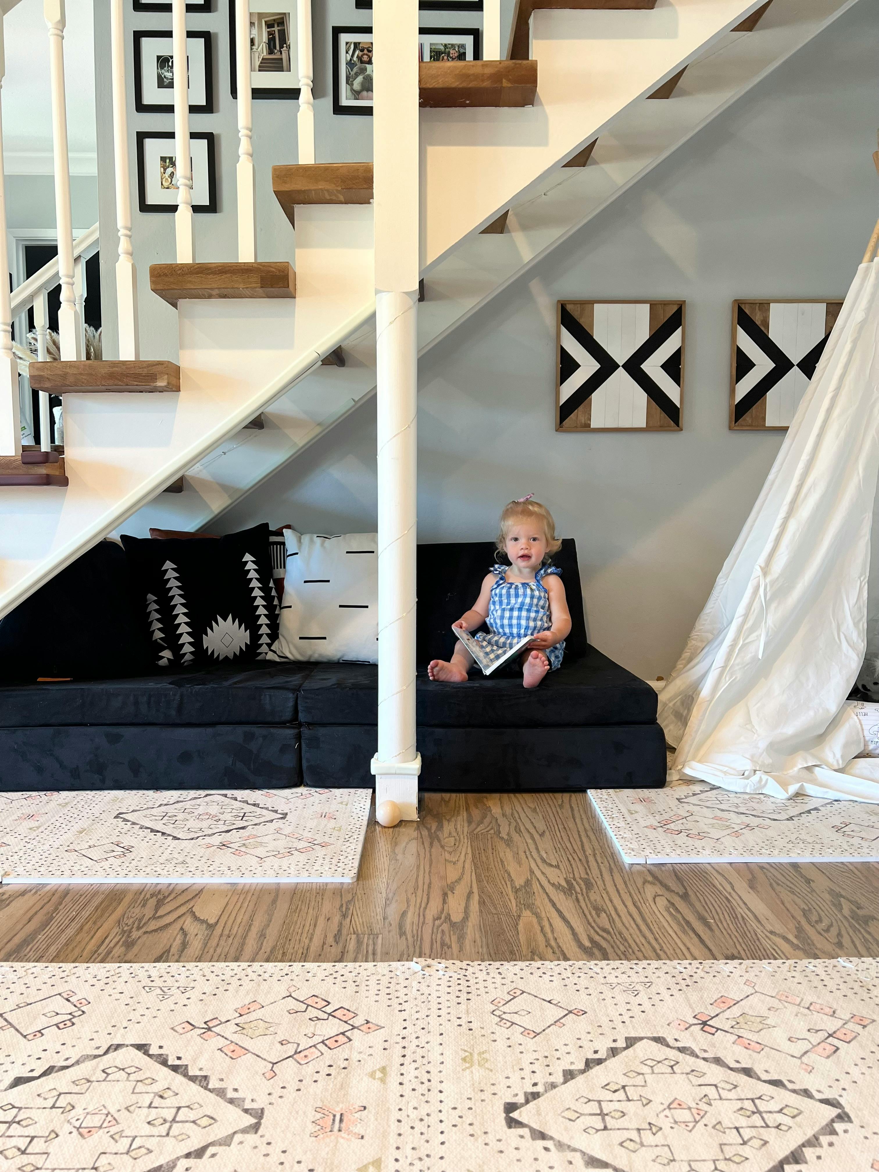 Child sitting on a black cushion under stairs, holding a book