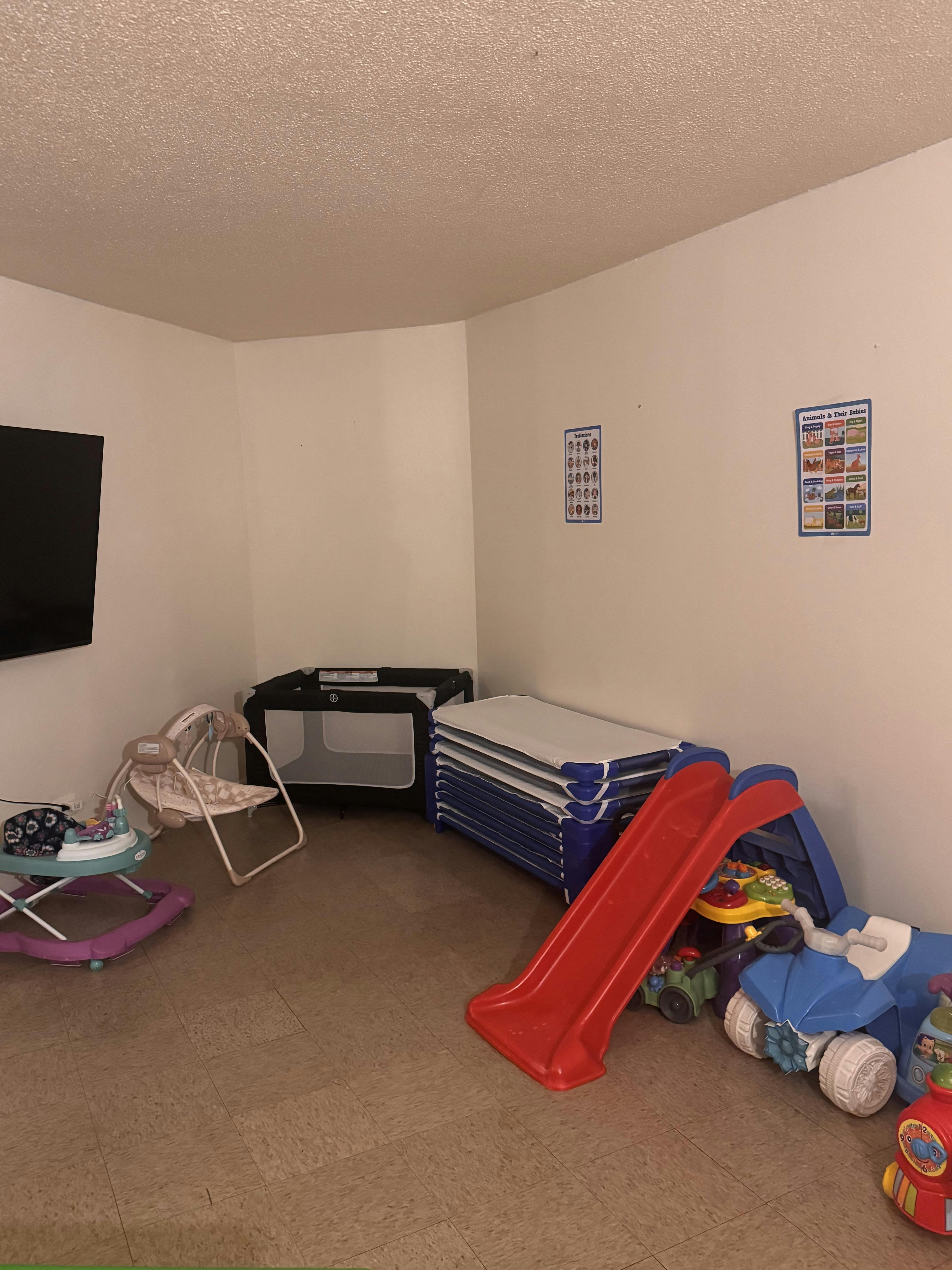 Childcare room with a crib, baby swing, walker, stack of cots, red slide, and toy car