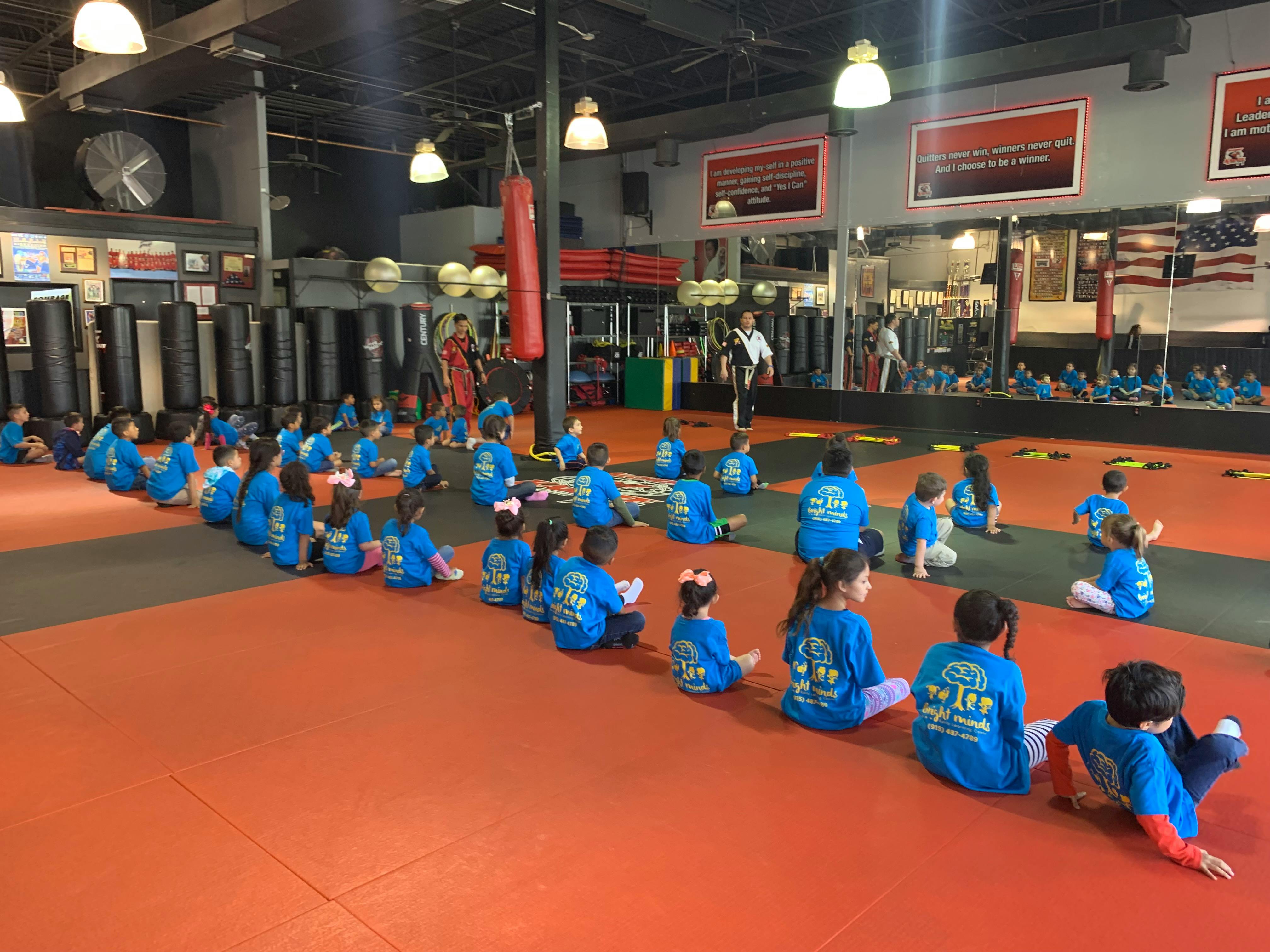 Group of children in blue shirts sitting on a training mat in a martial arts gym