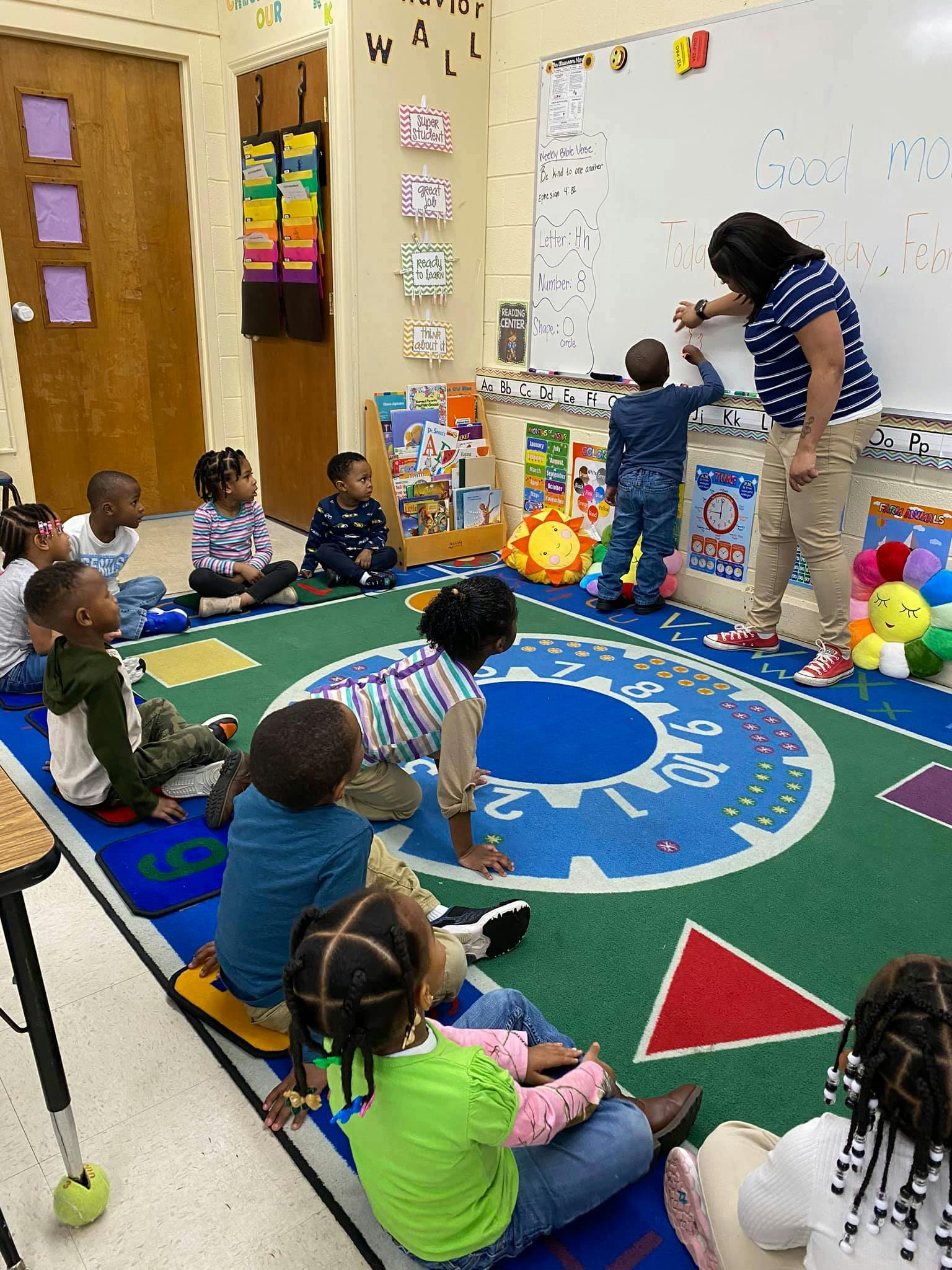 Adult and child writing on a whiteboard in a classroom with other children watching