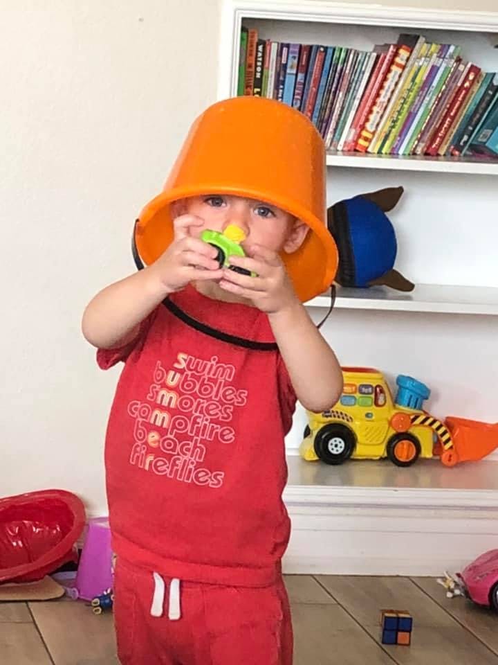 Child wearing an orange bucket on their head, holding a toy to their face