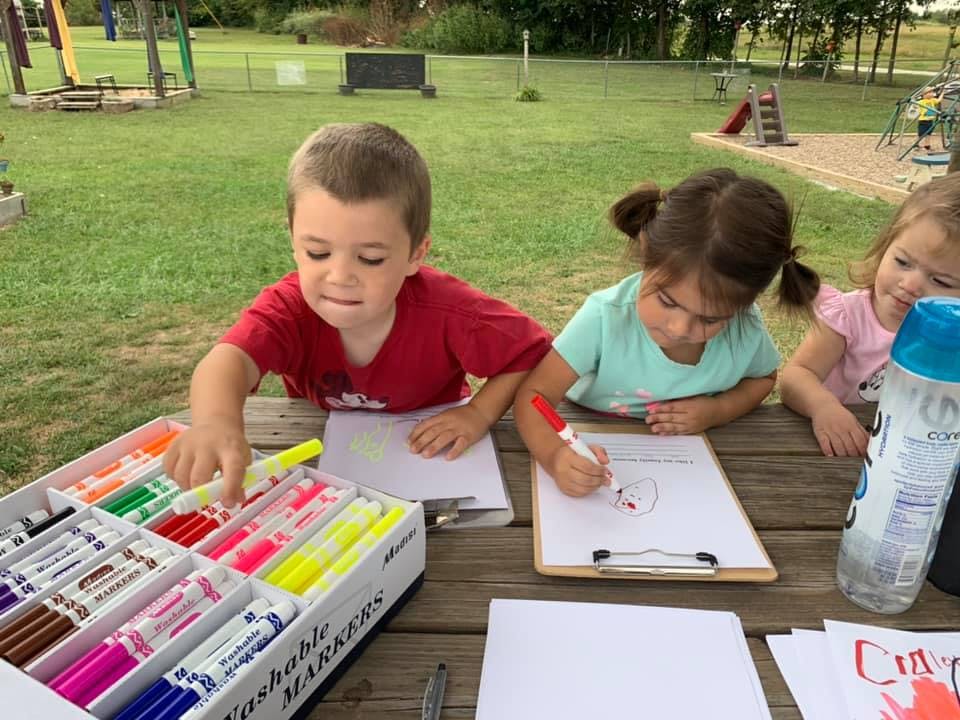 Three children drawing with markers at a picnic table outdoors