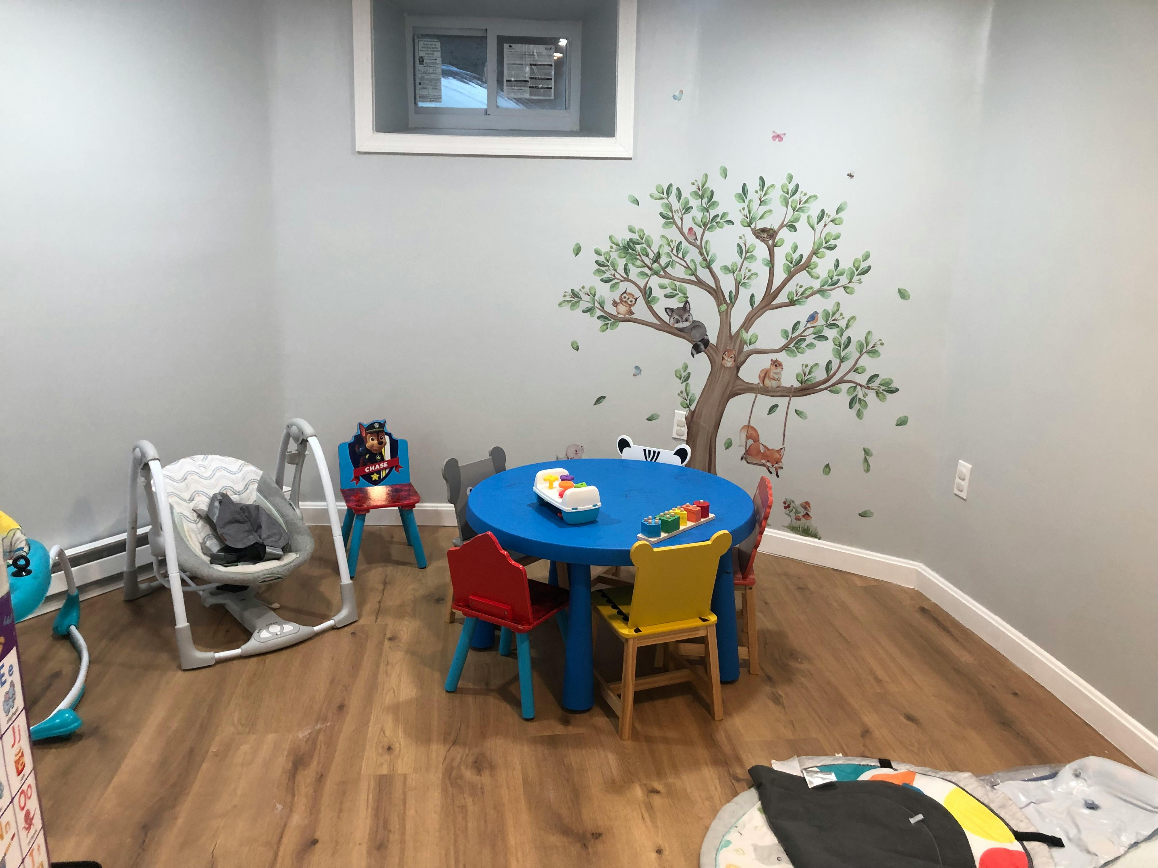 Playroom with a blue table and chairs, baby swing, and tree wall decal