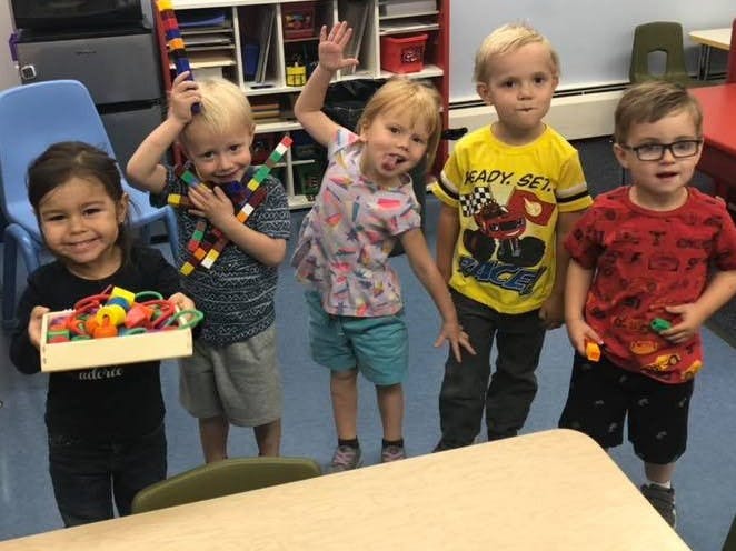 Five children standing in a classroom, one holding a box of toys