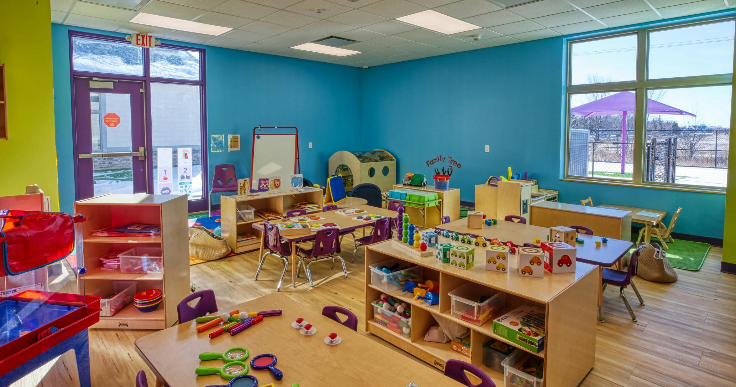 A colorful classroom with tables, chairs, and toys, an exit sign above a door