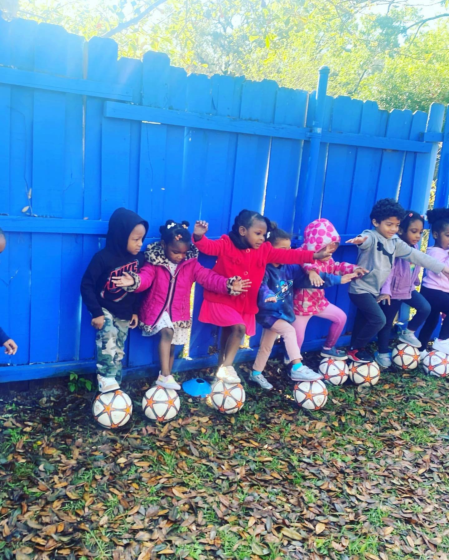 Children balancing on soccer balls against a blue fence