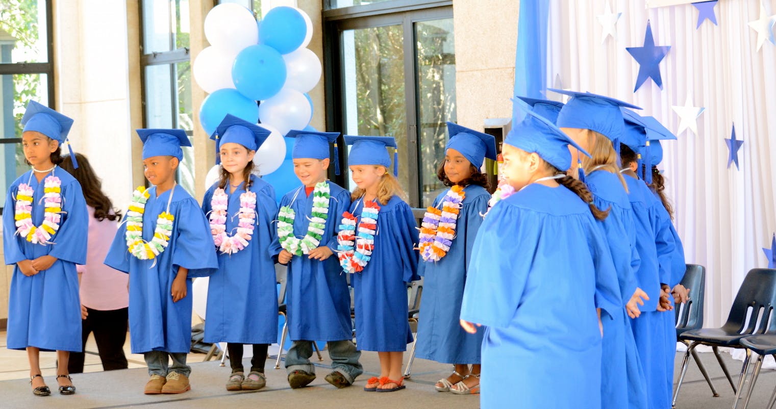 Children in blue graduation caps and gowns with leis standing on a stage