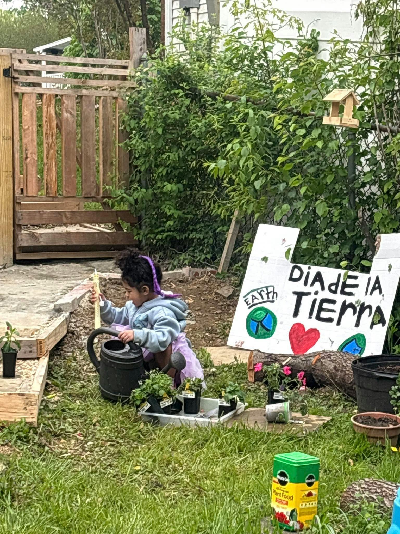 Child holding a stick over a watering can next to plants and a "Dia de la Tierra" sign