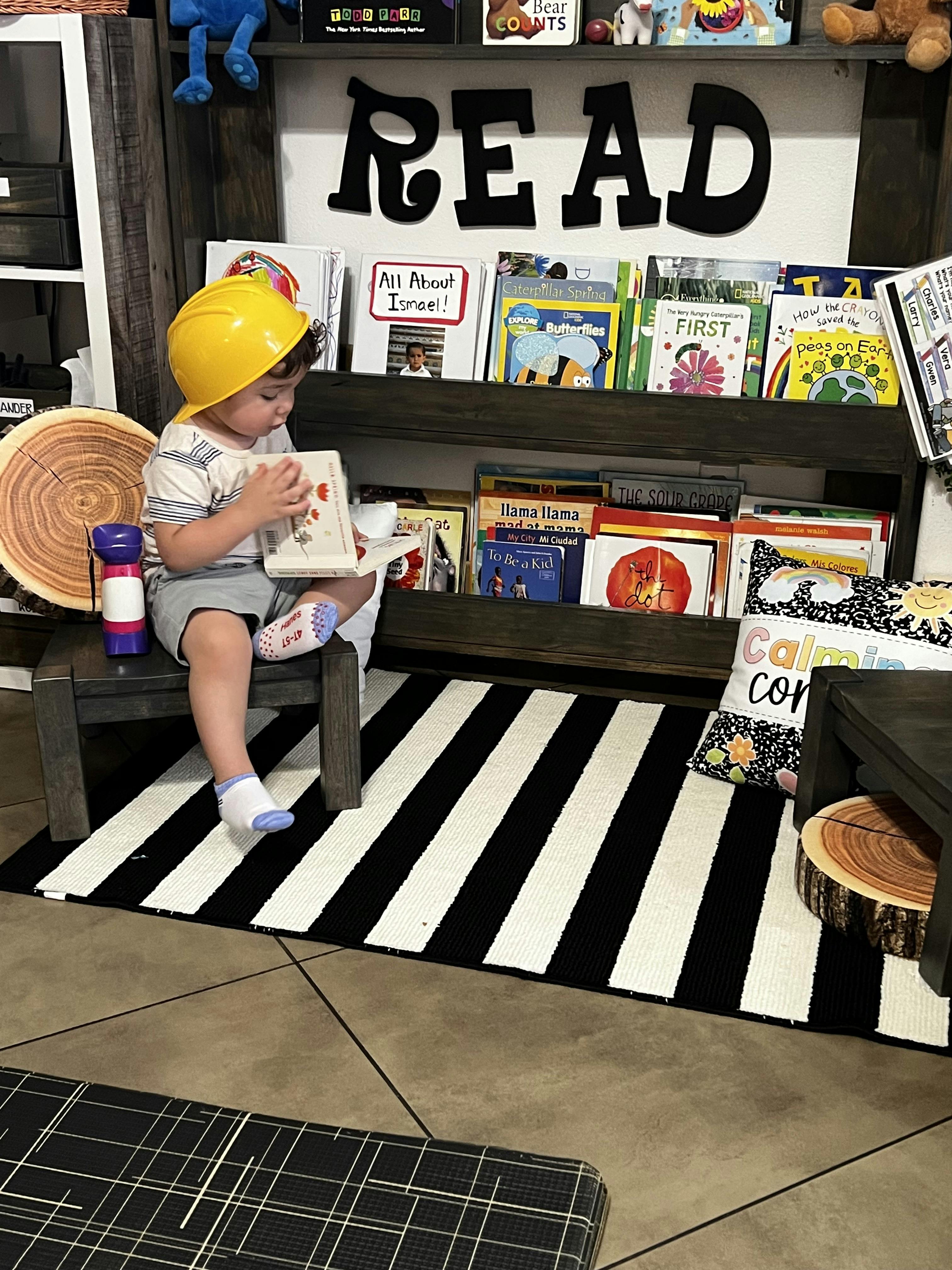 Child wearing a yellow hard hat reading a book in a library corner