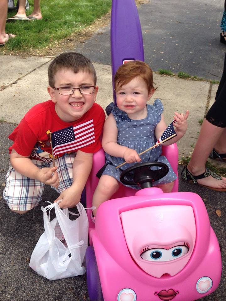 Child in glasses holding a flag next to a child in a toy car holding a flag