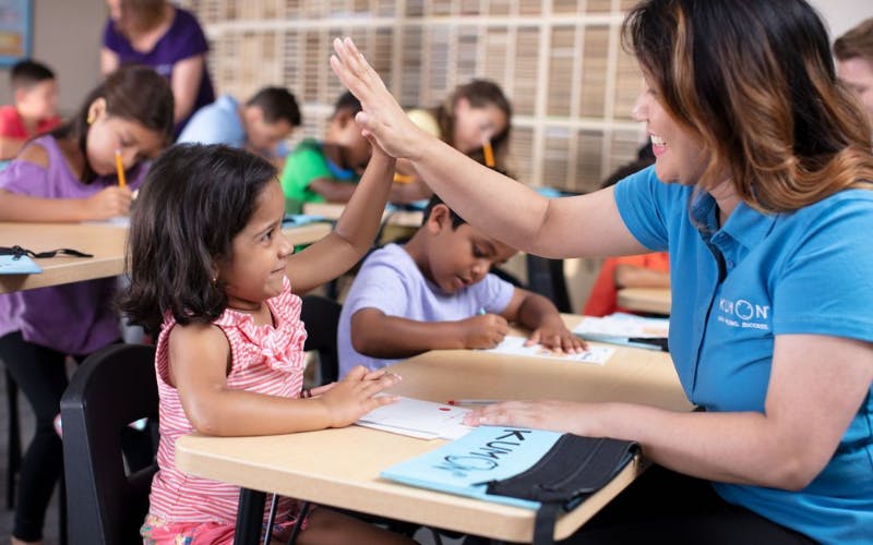 Adult giving a child a high-five in a classroom, other children working at desks