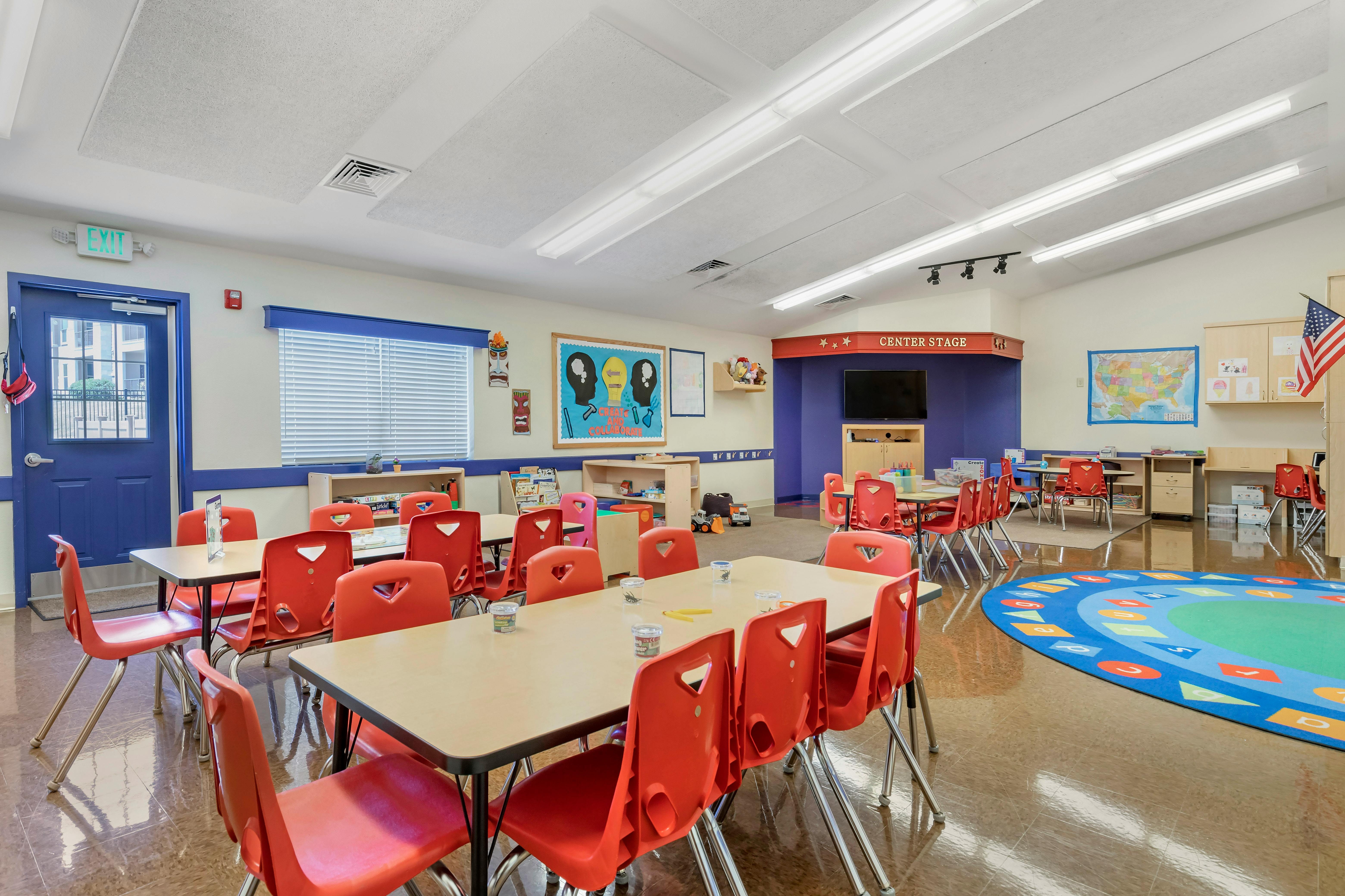 Classroom with small tables and red chairs, an alphabet rug, and a stage area