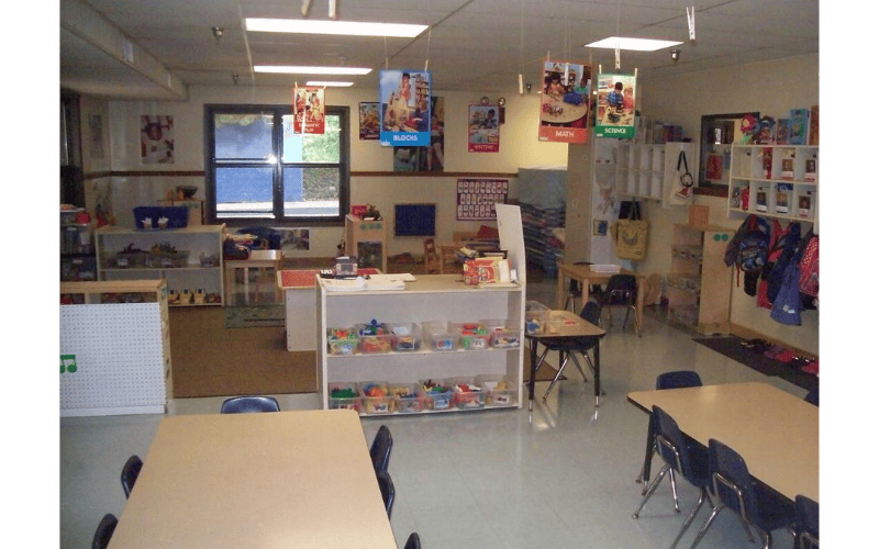 Empty classroom with small tables and chairs, shelves with toys, and posters