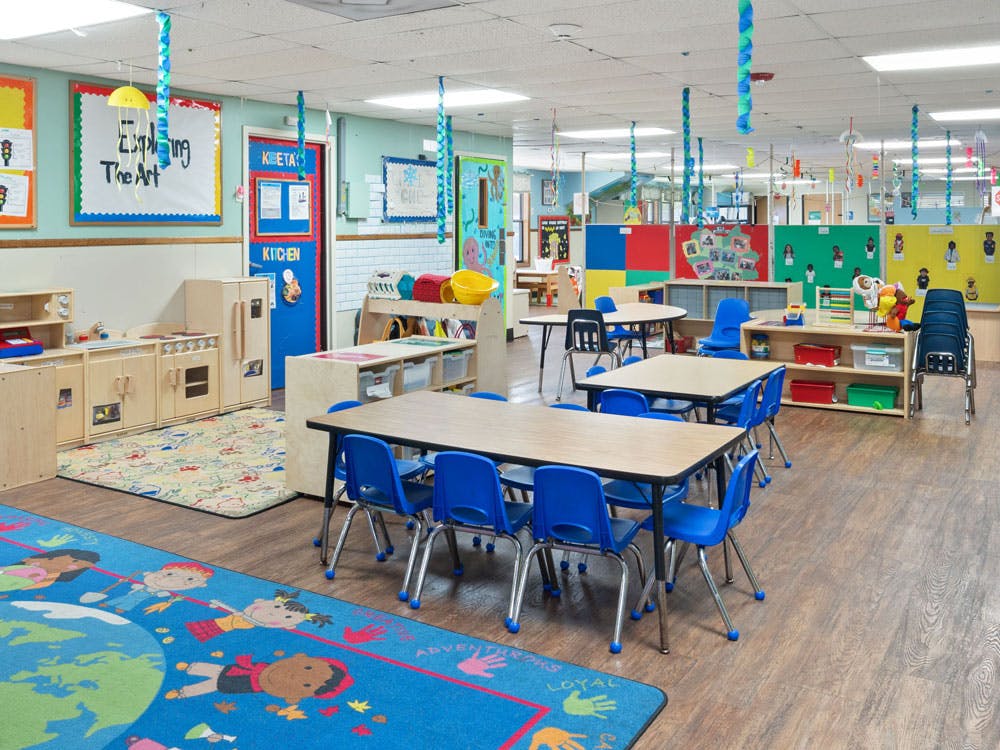 Colorful classroom with child-sized tables and chairs, play kitchen, and activity rug