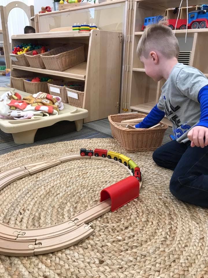Child playing with a wooden train set on a rug in a classroom