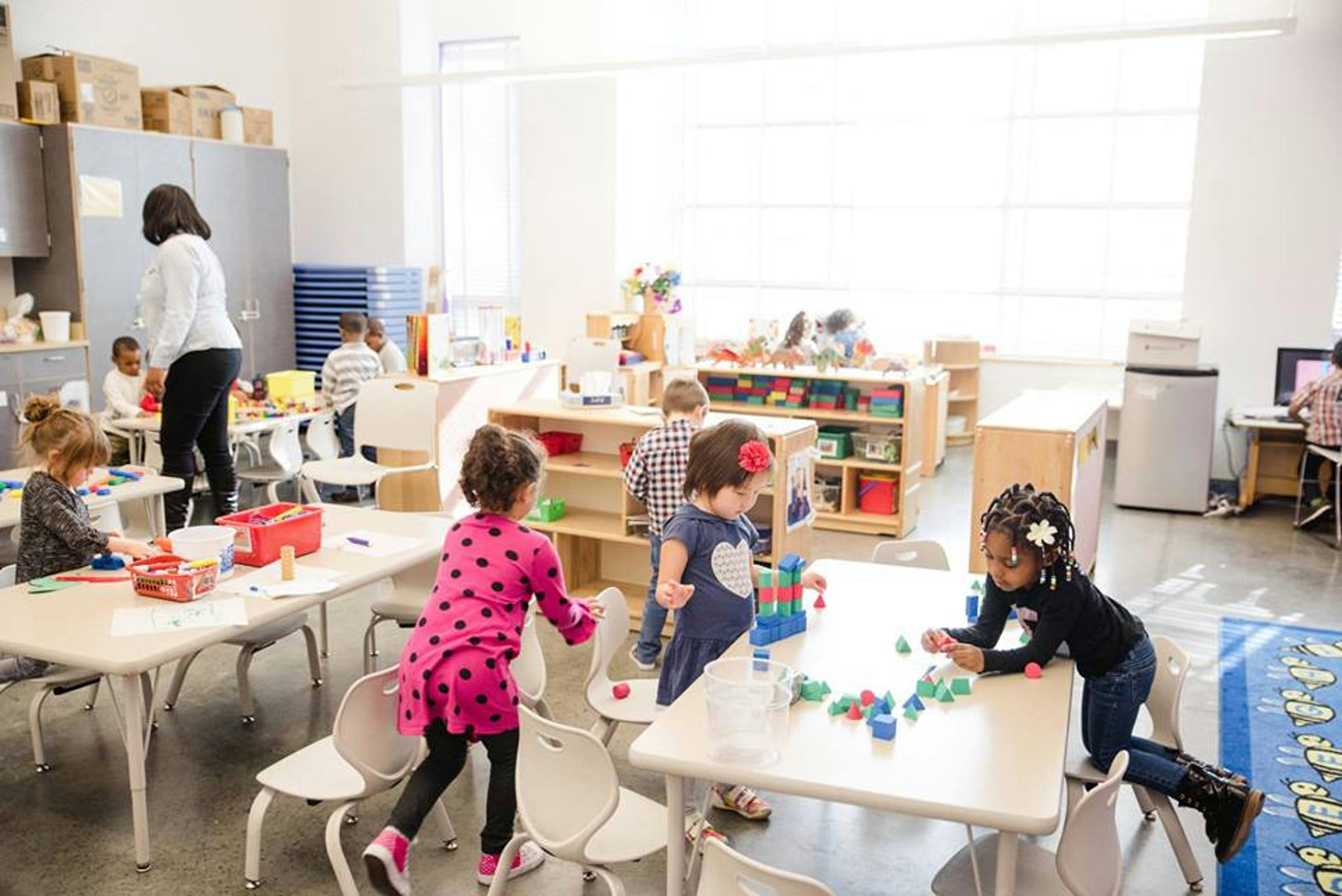 Children playing with toys at tables in a classroom with an adult supervising