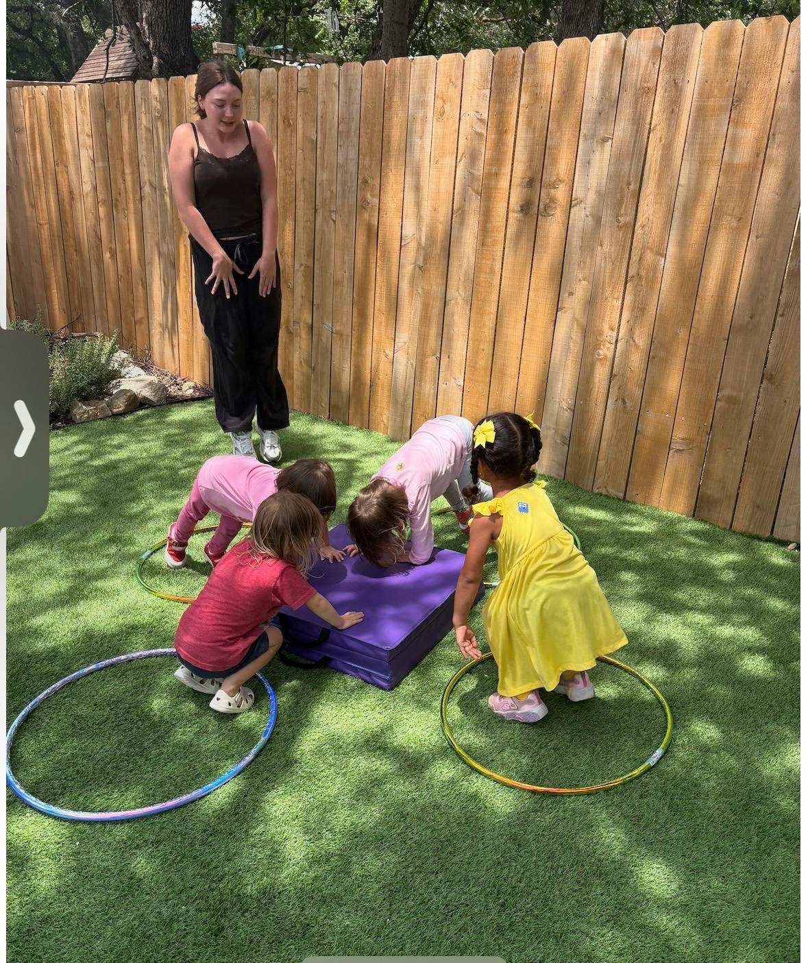 A person standing and watching four children around a purple mat on the grass