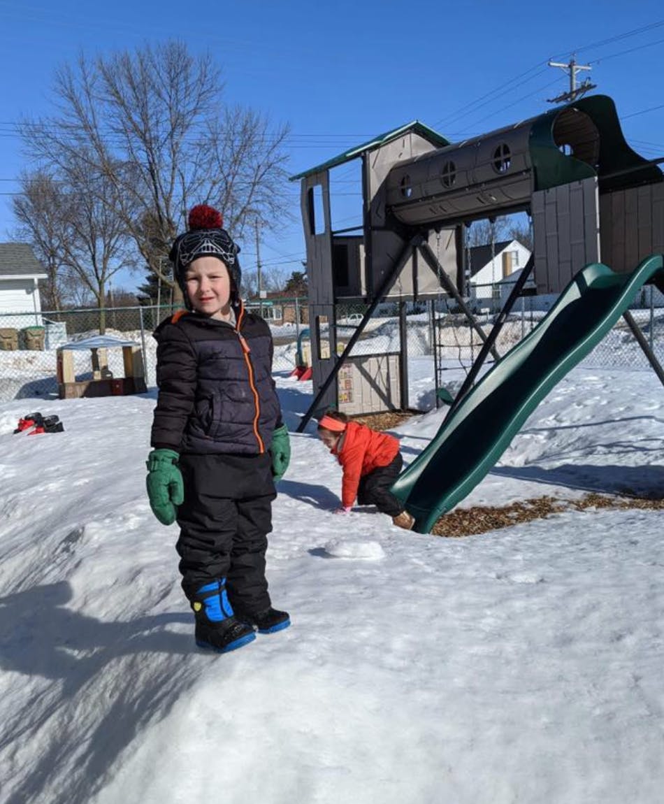 Child standing in snow next to a playground slide, another child at the bottom