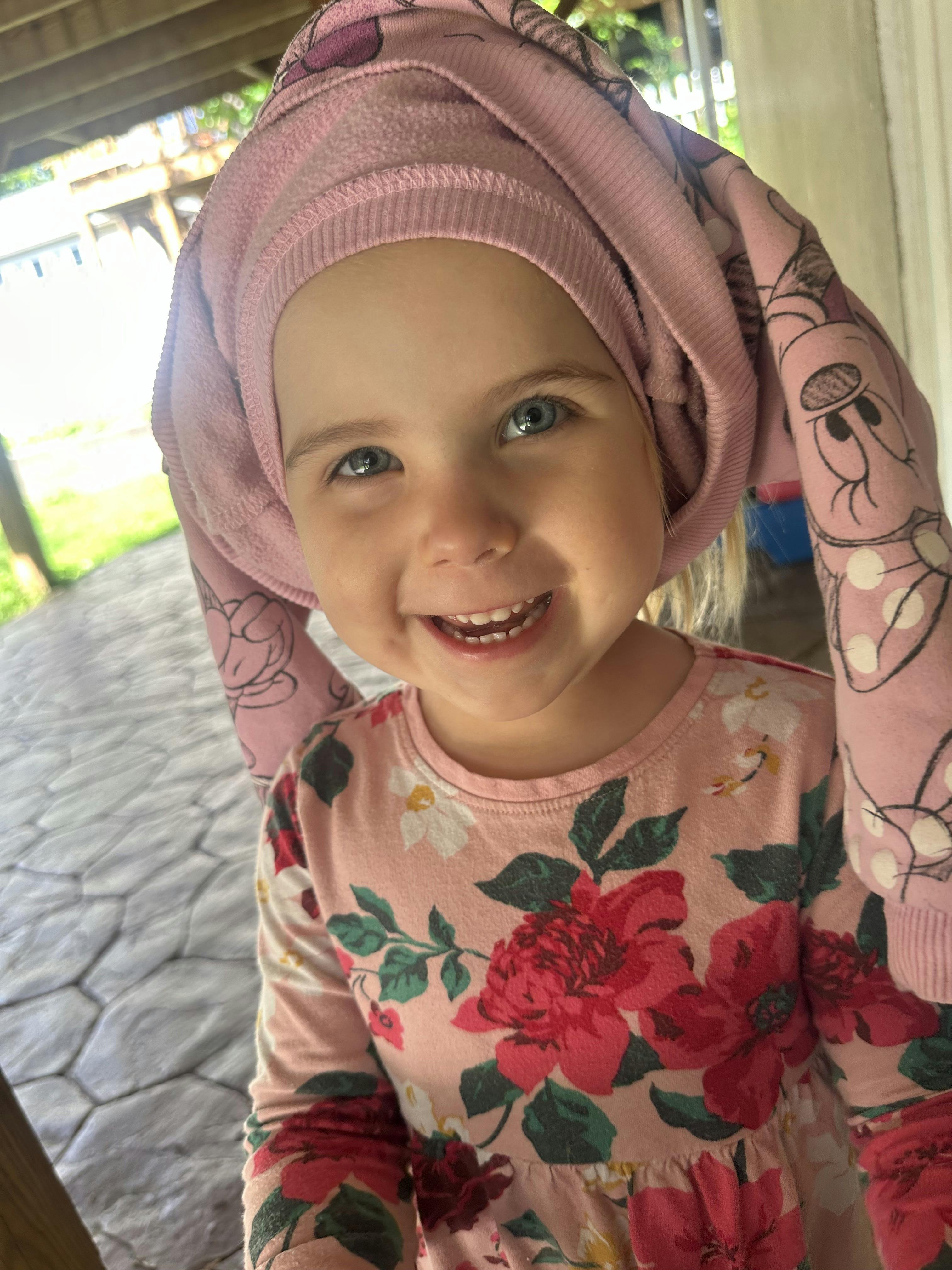 Child smiling with a pink hooded sweatshirt wrapped around their head