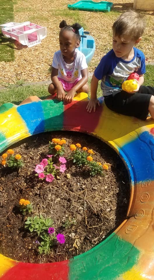 Two children sitting by a colorful tire planter with flowers