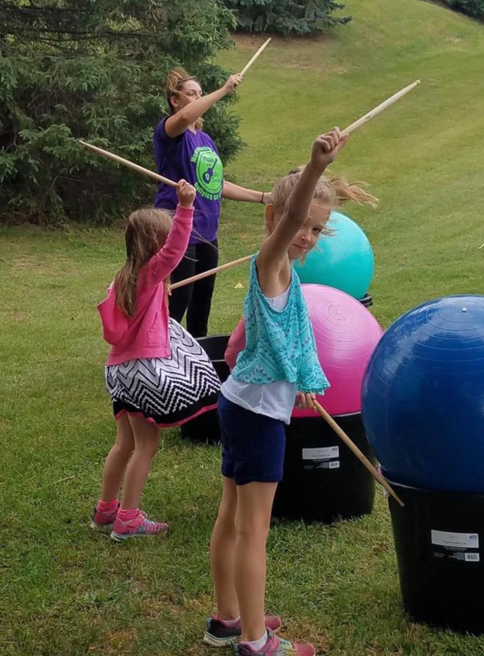 Two children and an adult playing drums outdoors with drumsticks