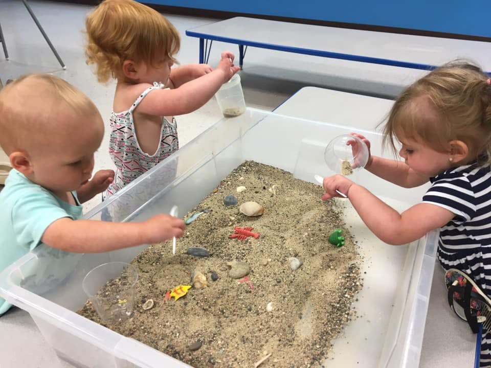 Three children playing with sand and small objects in a large bin