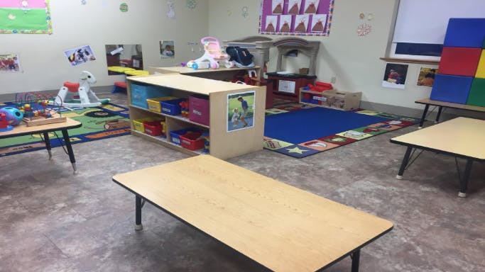 Empty classroom with small tables, rugs, and shelves with toys