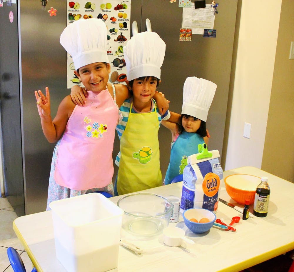 Three children in chef hats and aprons standing behind a table with baking ingredients