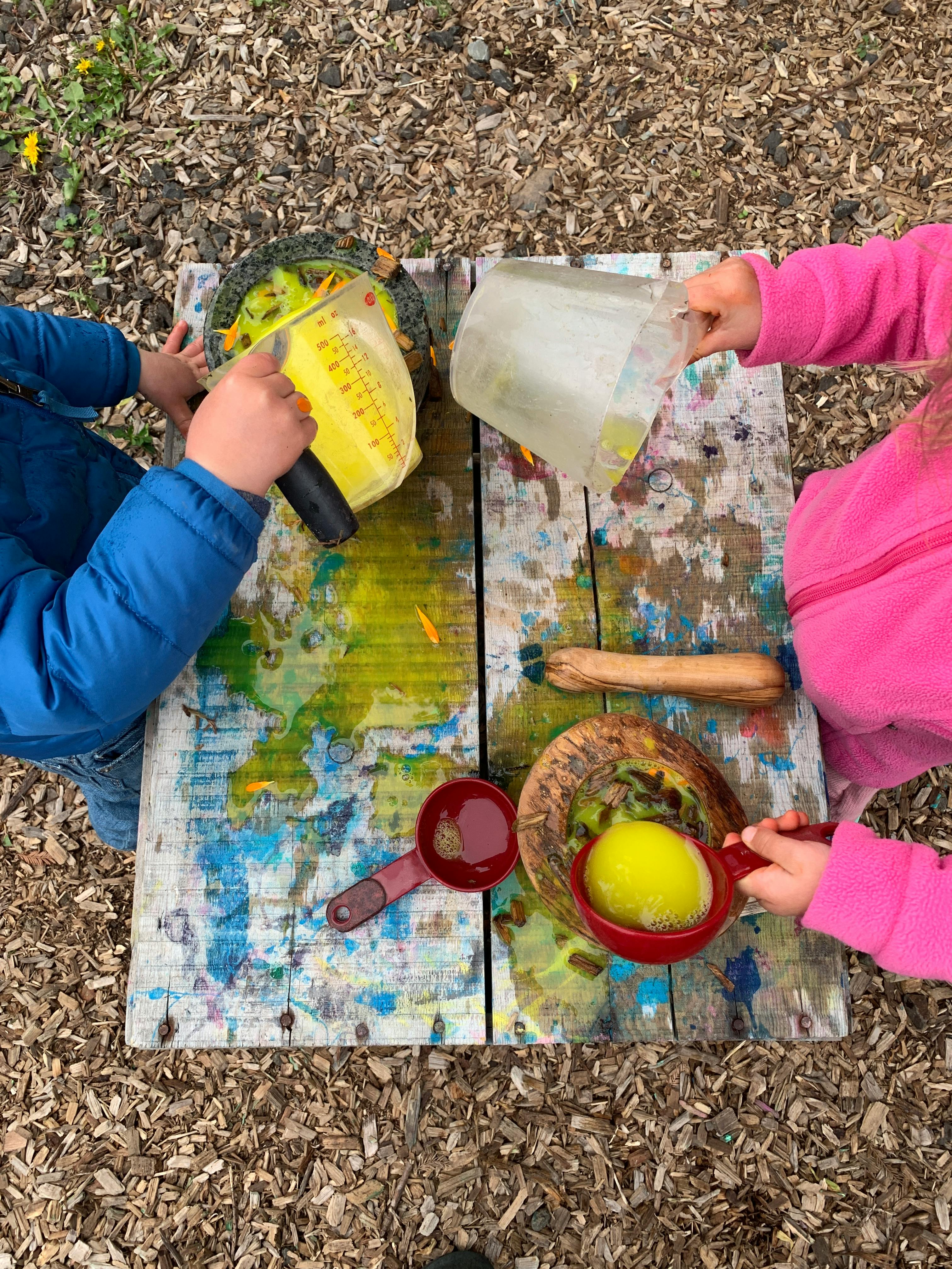 Two children playing with various liquids and natural materials on a wooden surface