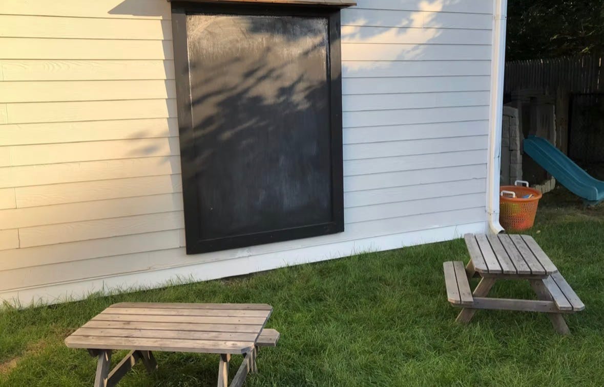 Chalkboard on a wall with two wooden picnic tables on grass