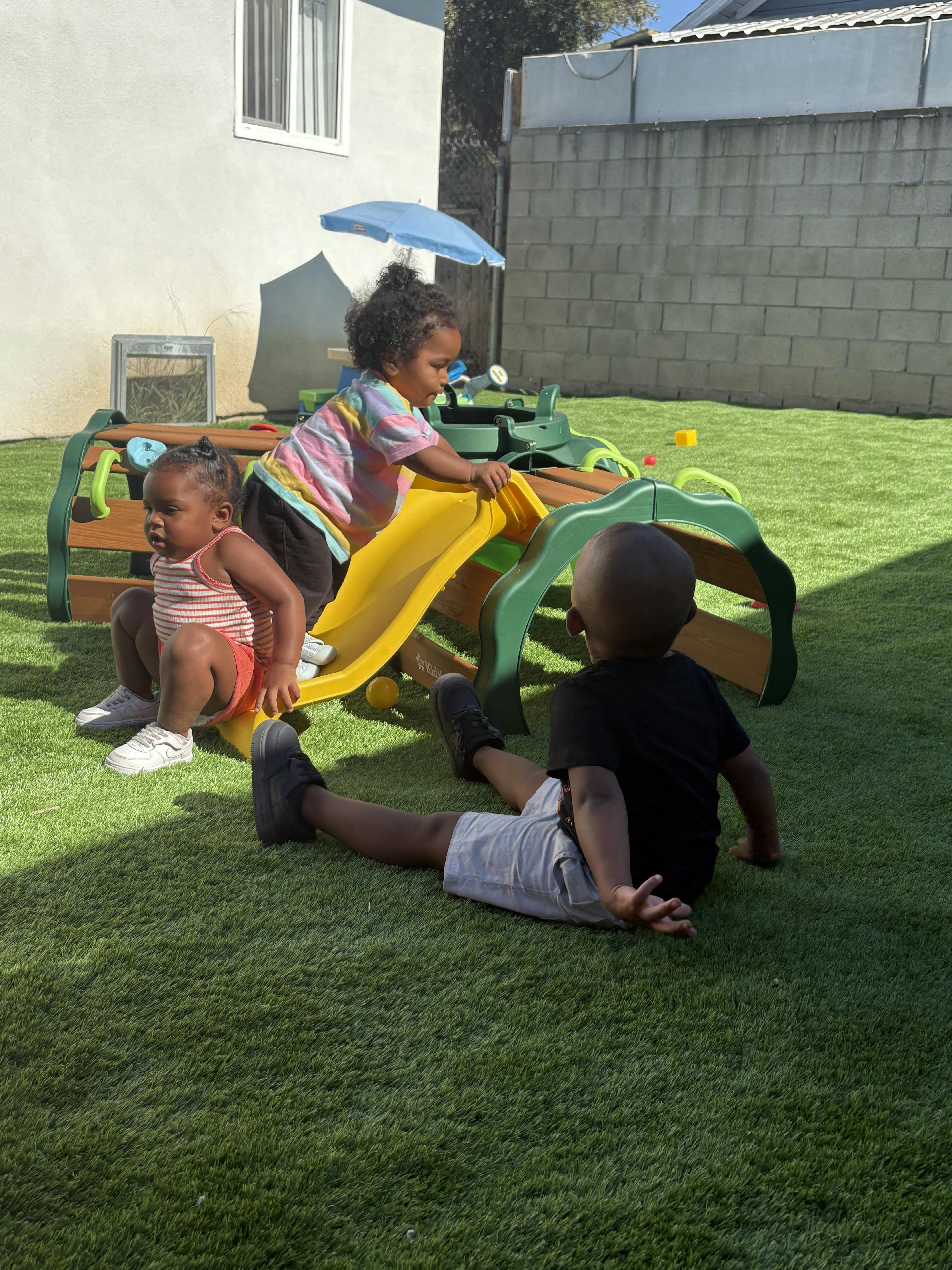 Three children playing on a small slide in a grassy area