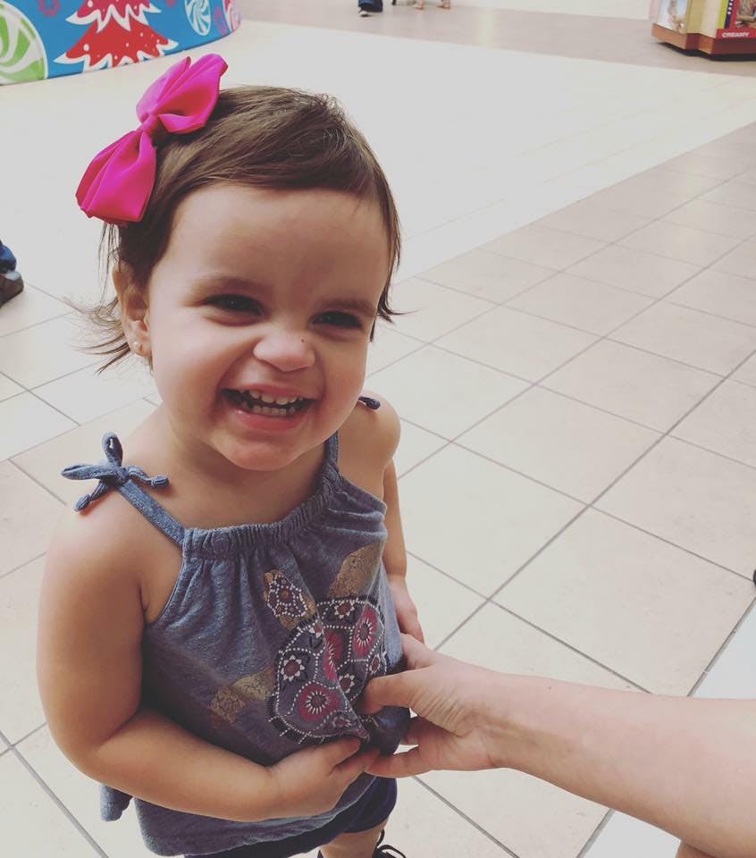 Child with a pink bow in hair, smiling, holding a person's hand