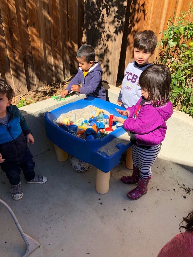Three children playing with sand and toys at a blue activity table