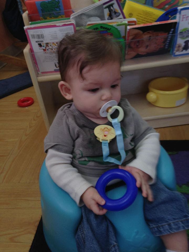 Child with a pacifier in a blue chair holding a blue ring