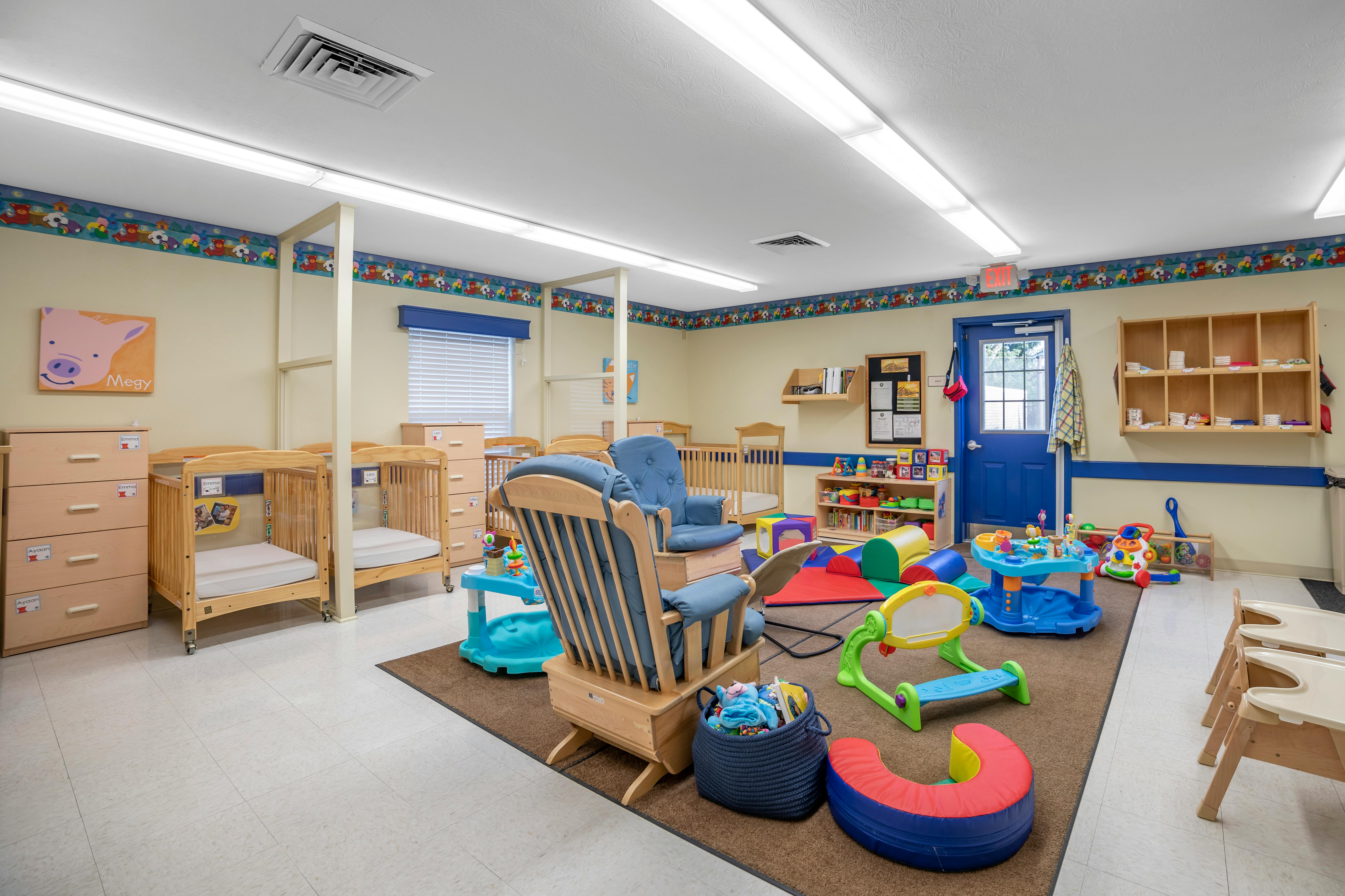 A colorful childcare classroom with cribs, play mats, toys, and small furniture