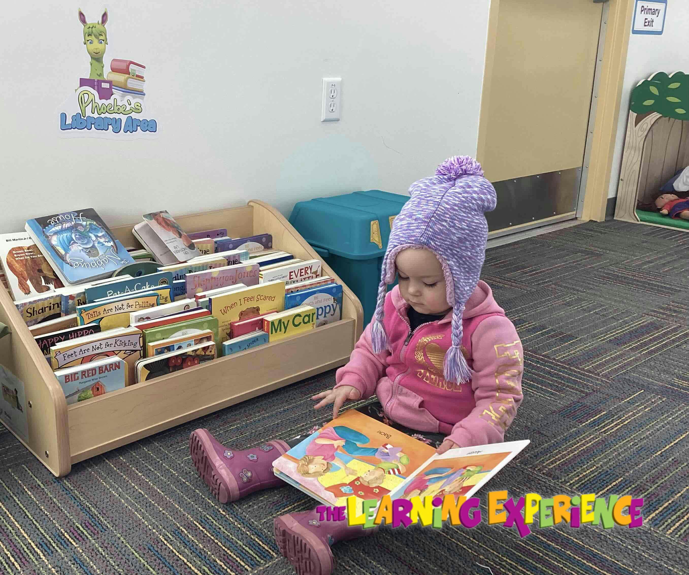 Child in a purple hat sitting on the floor reading a book next to a bookshelf