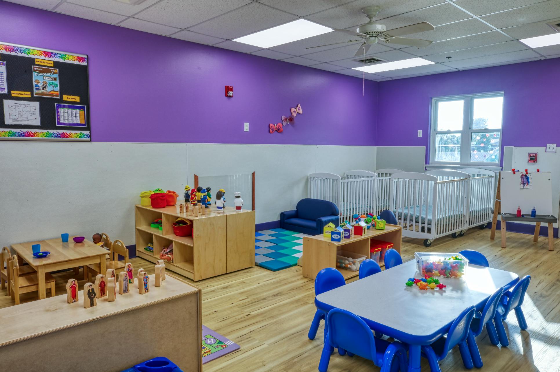 Classroom with small tables and chairs, cubbies with toys, and three cribs