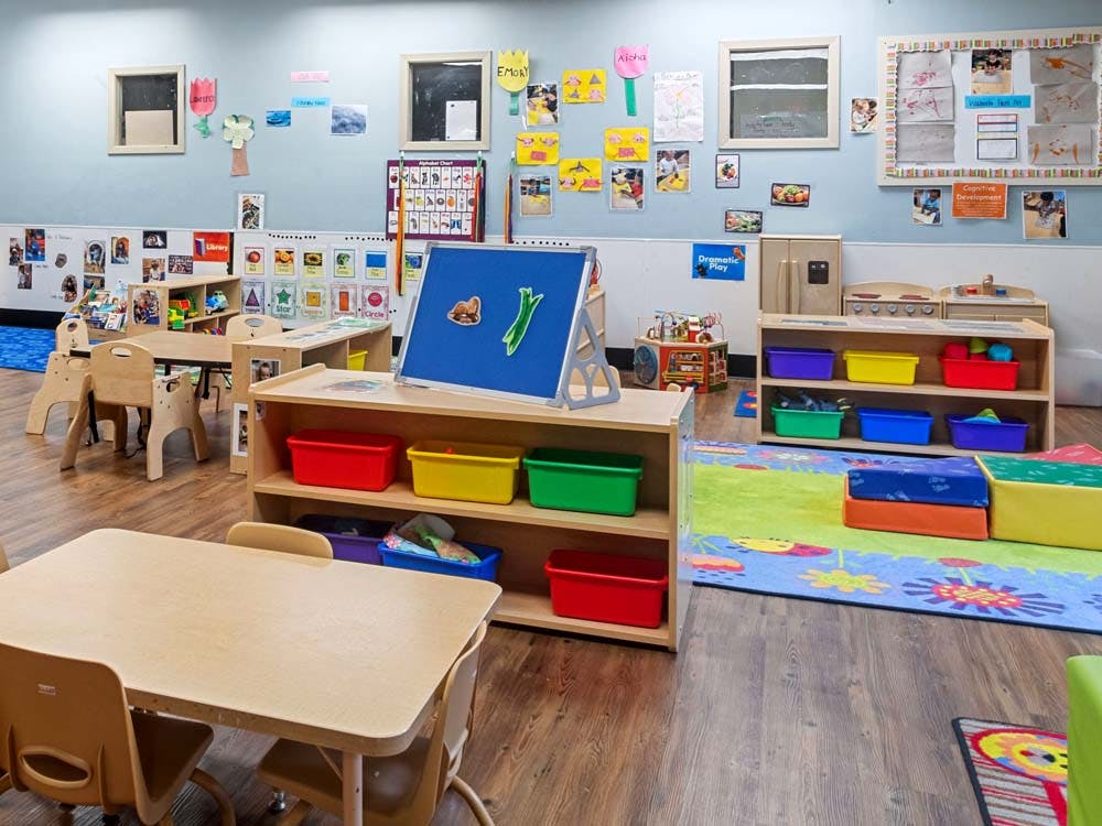A classroom with small tables and chairs, shelves with colorful bins, and play kitchen items