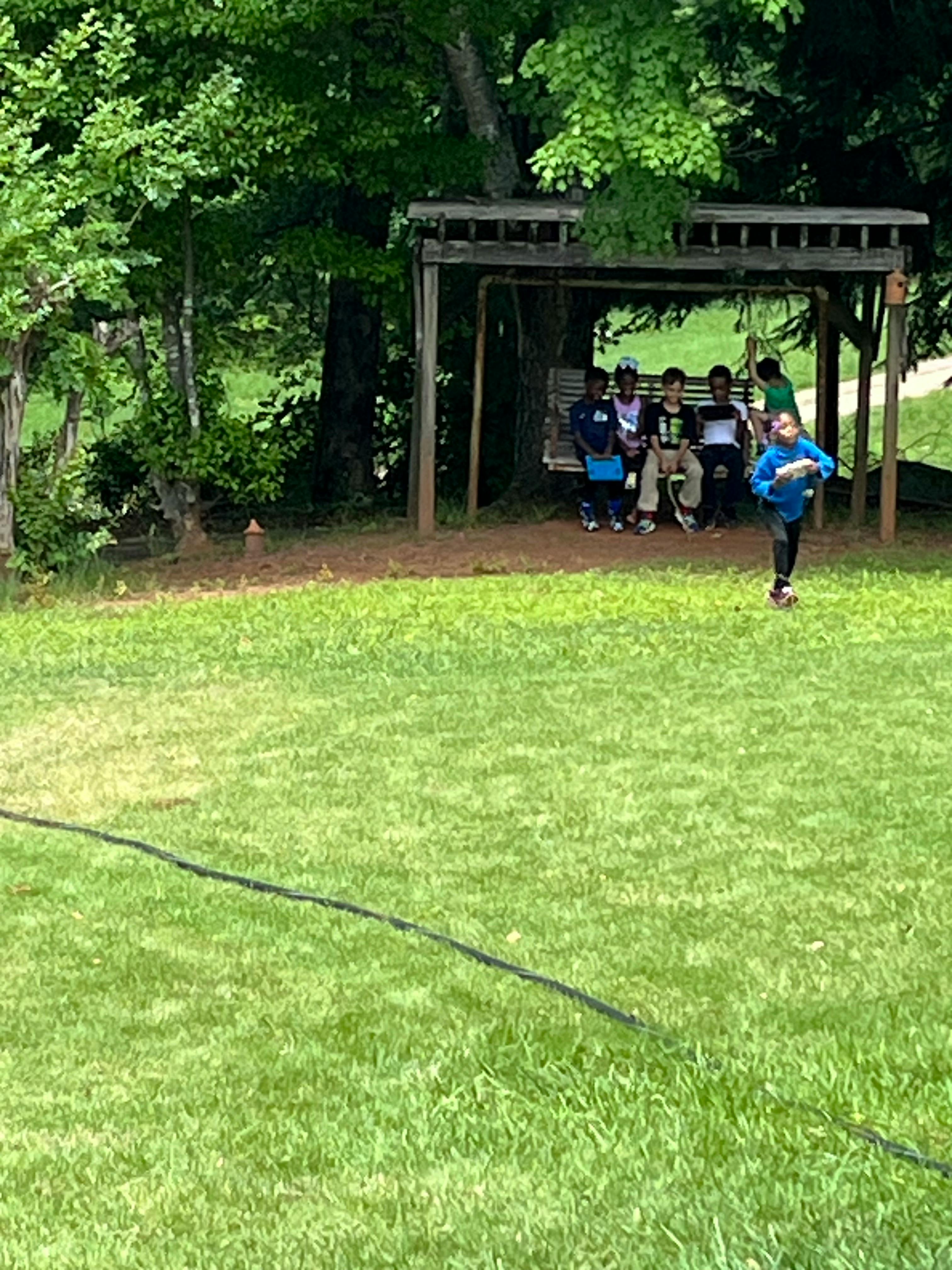 Child running in grass with other children sitting on a swing in the background