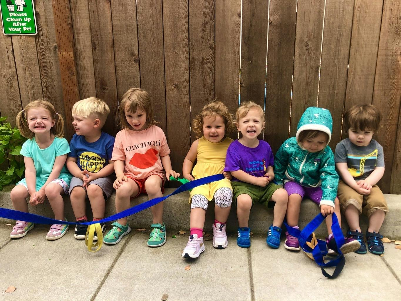 Six children sitting in a row against a wooden fence