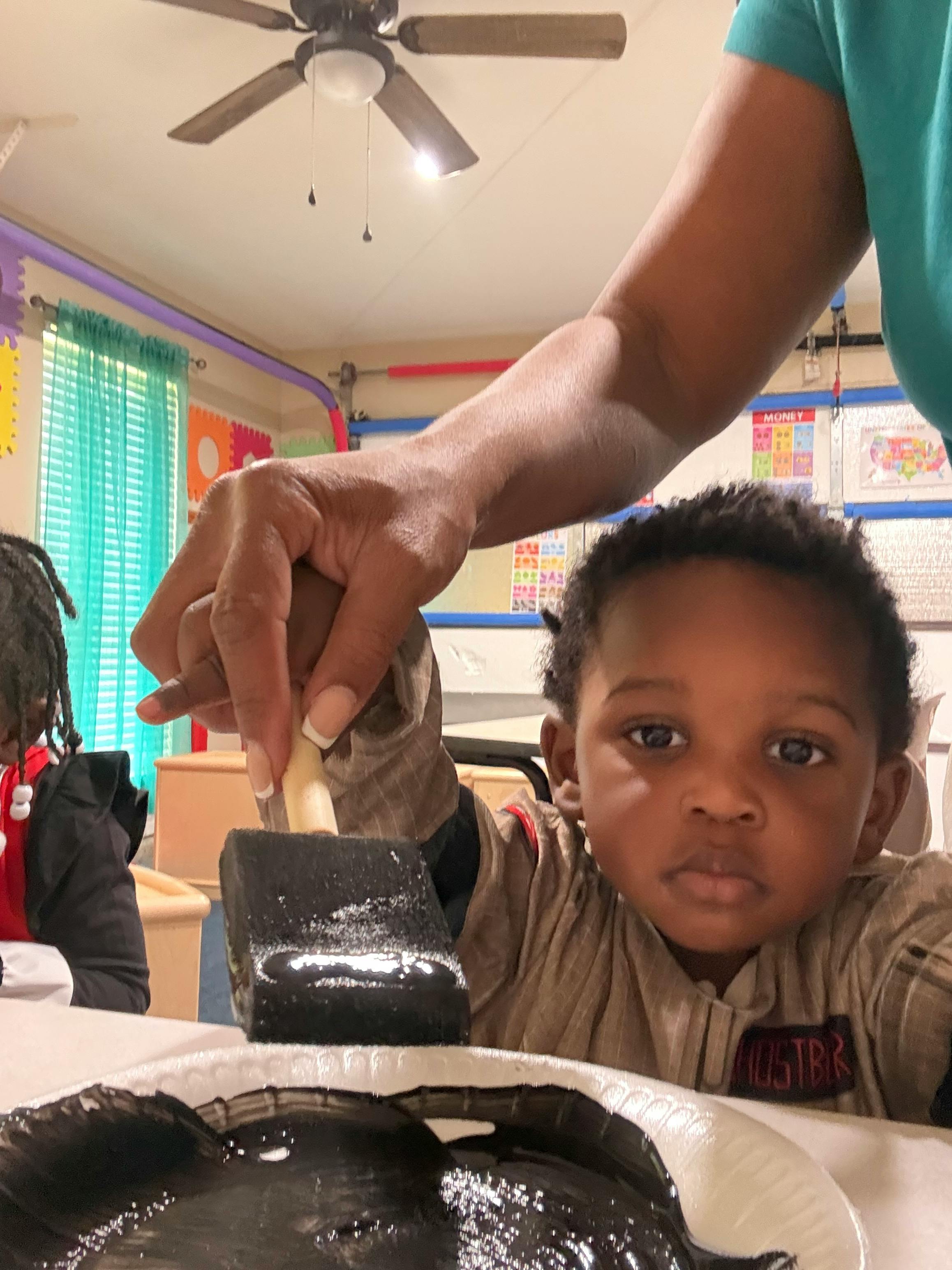 Adult hand helping a child paint with a brush and black paint