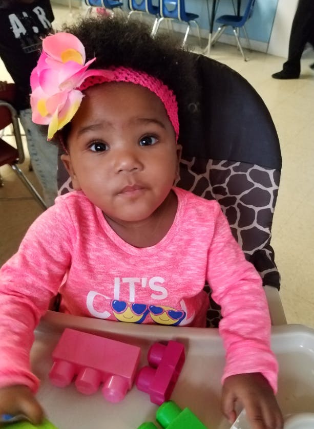 Child in a high chair with a pink headband and flower, "It's Cool" on shirt
