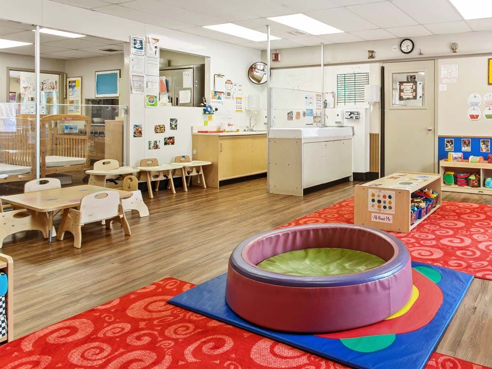 Empty infant classroom with cribs, small tables, and play mats